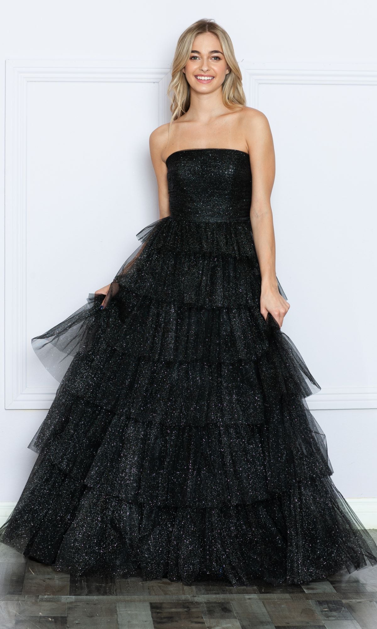 Smiling woman in the Strapless Long Tiered Glitter Prom Ball Gown 9386 in black, featuring a glittery tulle overlay and tiered skirt, stands against a white wall.